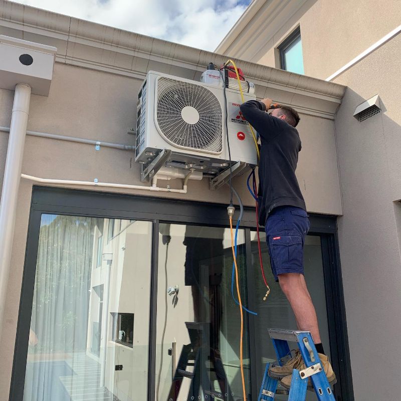 Technician on a ladder working on an outdoor unit mounted above glass doors