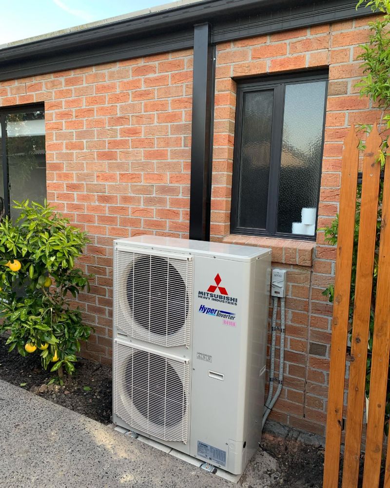 Large two-fan outdoor air conditioning unit beside a brick house and fruit tree