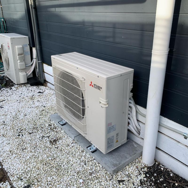 Two outdoor air conditioning units on white pebbles beside a dark weatherboard wall