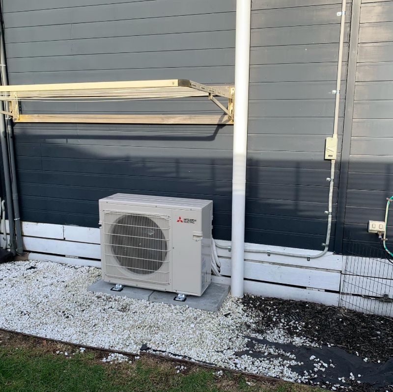 Outdoor air conditioning unit on a pebble bed beside a dark weatherboard house